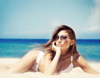 woman in swimsuit and sunglasses lounging on stomach on beach with chin in hand in front of ocean and sky