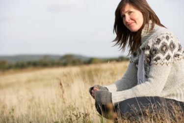 smiling woman in gray long sleeve and dark pants sitting in a field in front of cloudy sky